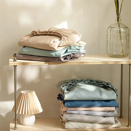 Stacks of folded clothes on a wooden shelf with a lamp and vase in the background.