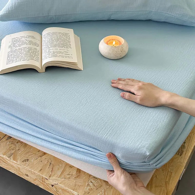 Light blue mattress cover on a bed with an open book and candle, hand touching the fabric.