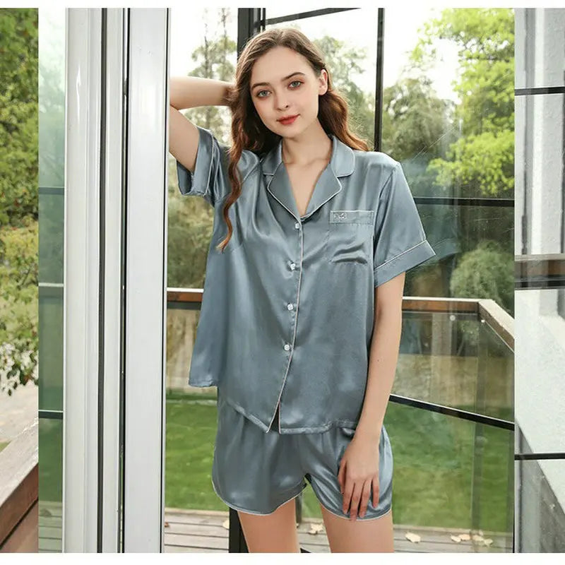 Woman wearing a light blue satin pajama set standing in front of a glass door with greenery outside.