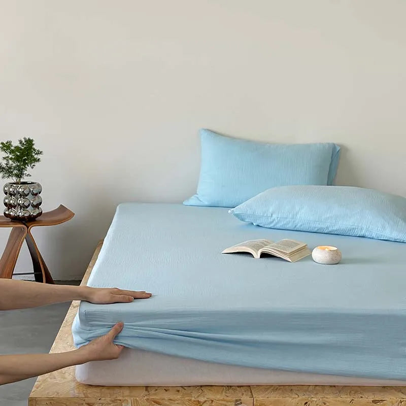 Person adjusting light blue fitted sheet on a bed with pillows and a plant in the background.