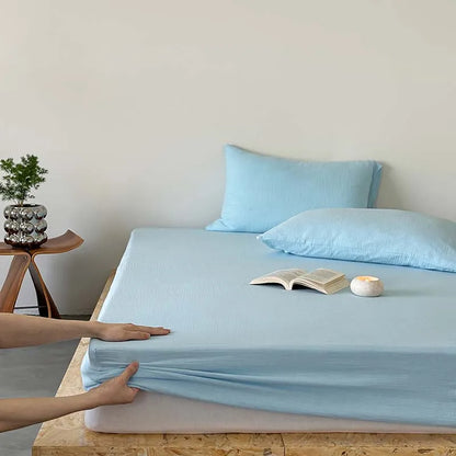 Person adjusting light blue fitted sheet on a bed with pillows and a plant in the background.