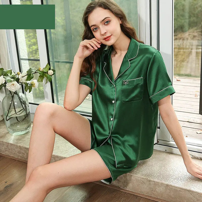 Woman wearing a green satin pajama set sitting by a window with a vase of flowers.