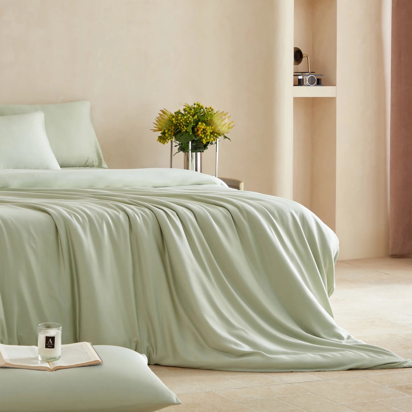 Light green bedspread on a bed in a softly lit room with flowers and a candle.