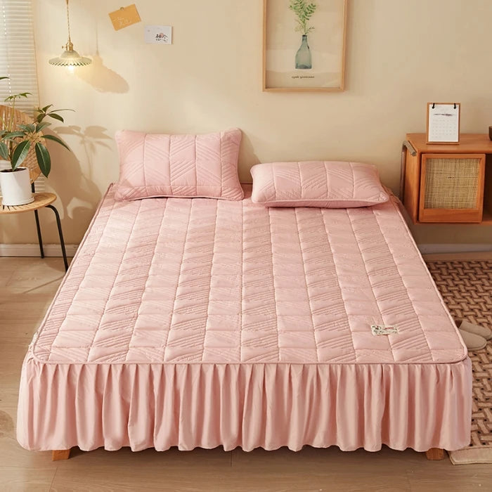 Pink quilted bedspread on a bed in a room with a lamp, plant, and side table.