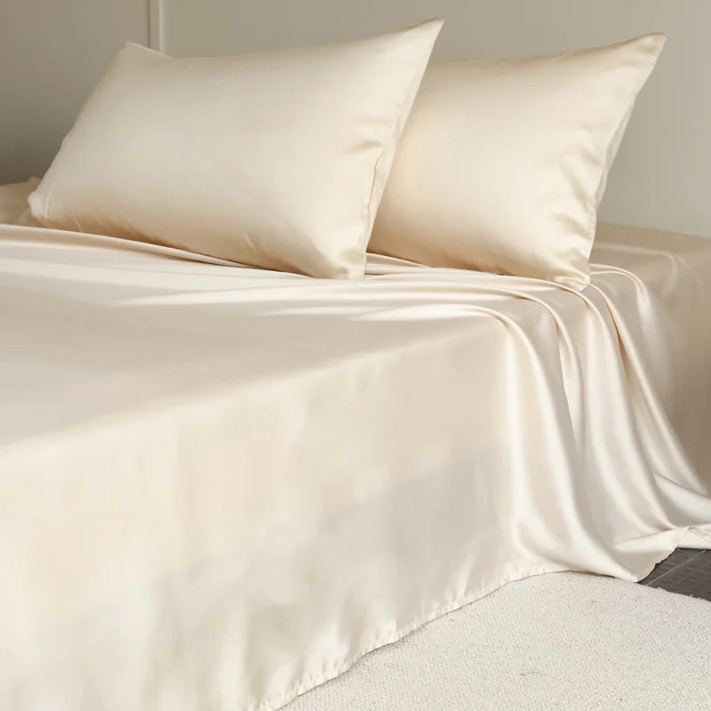 Beige satin sheet set on a bed with pillows against a neutral background
