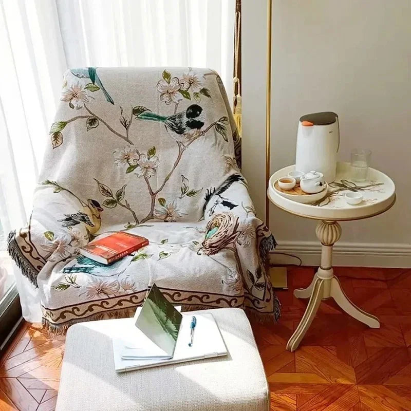 Decorative armchair with floral and bird pattern, next to a small round table with a lamp and books.