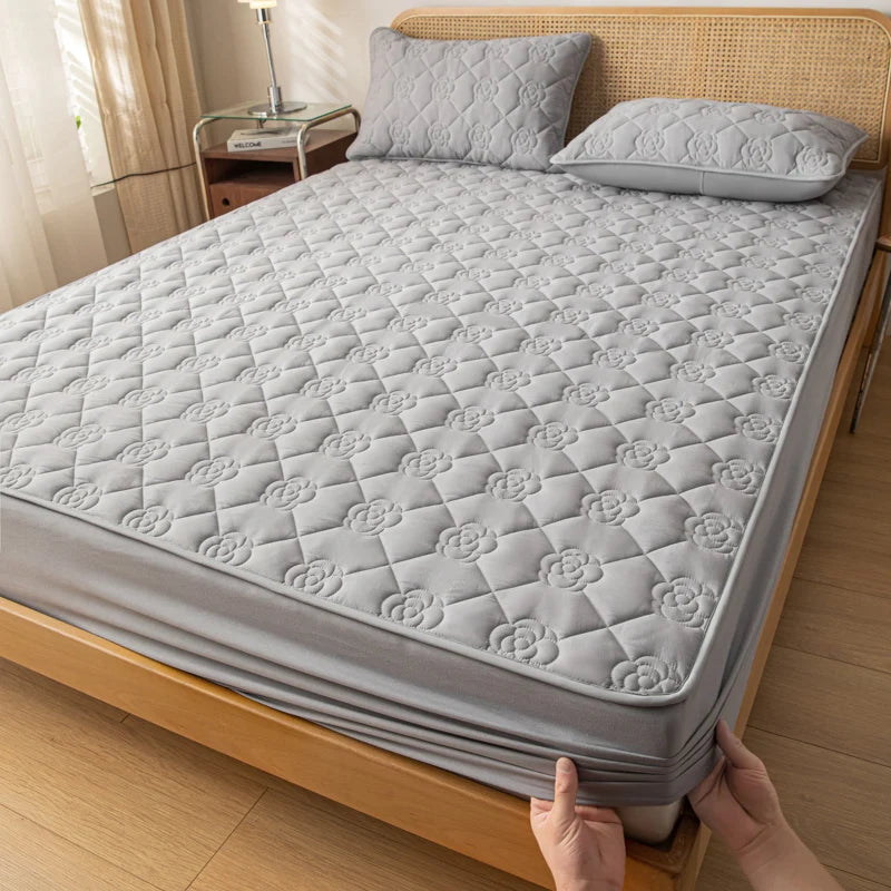 Gray quilted mattress cover being fitted onto a bed in a bedroom setting.