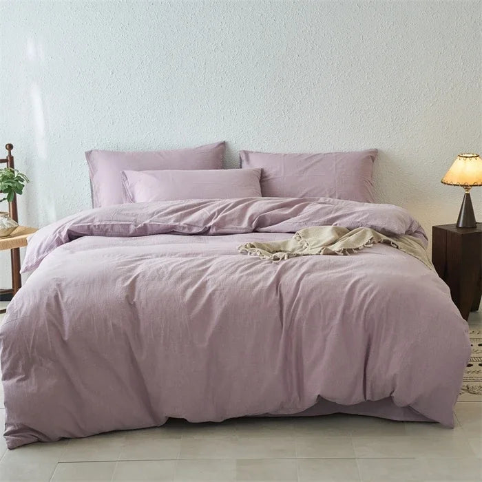 Bed with purple bedding in a room with a lamp and plant.