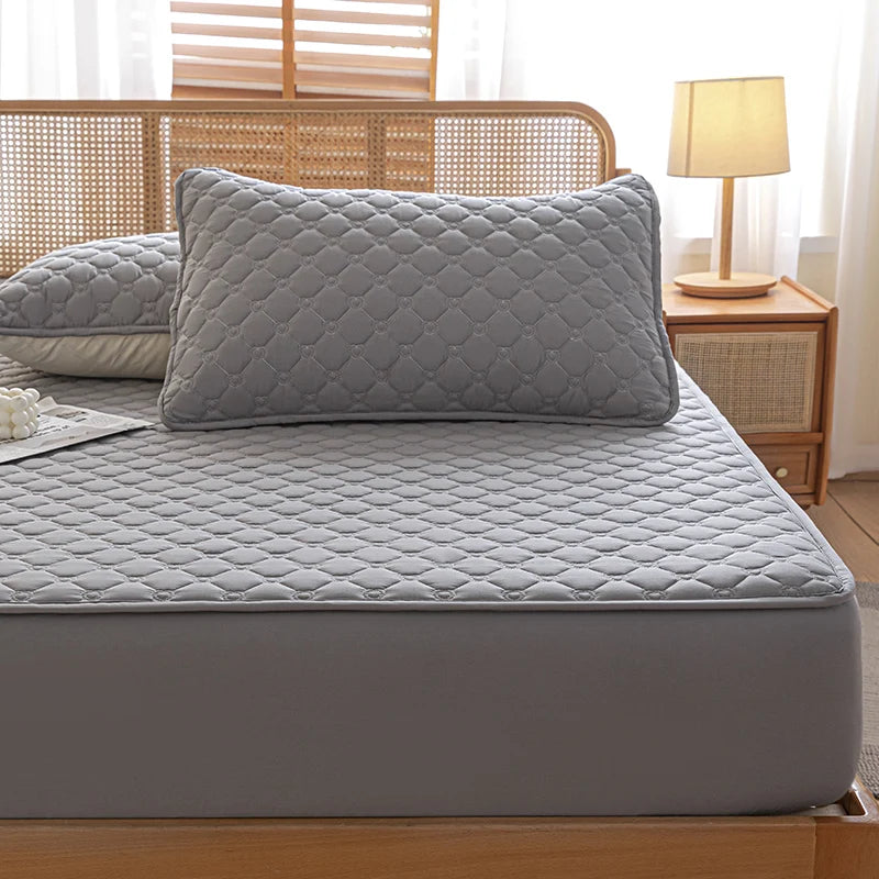 Gray quilted mattress cover on a bed with pillows and a lamp in the background.