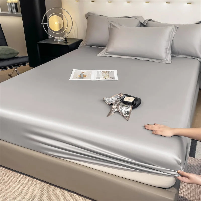 Person adjusting a silver satin sheet on a bed with a nightstand and lamp in the background.