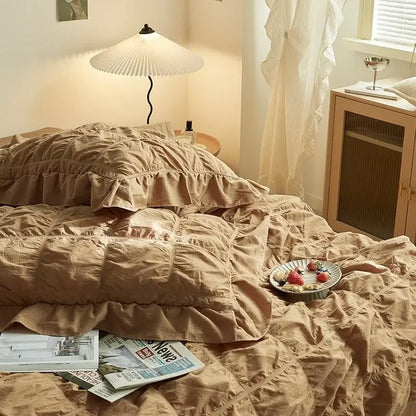 Bed with beige comforter and pillows, side table with lamp, and window with white curtains.