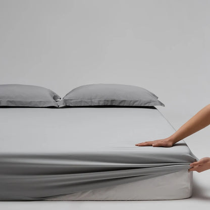 Person unrolling a gray fitted sheet on a bed with gray pillows against a plain background