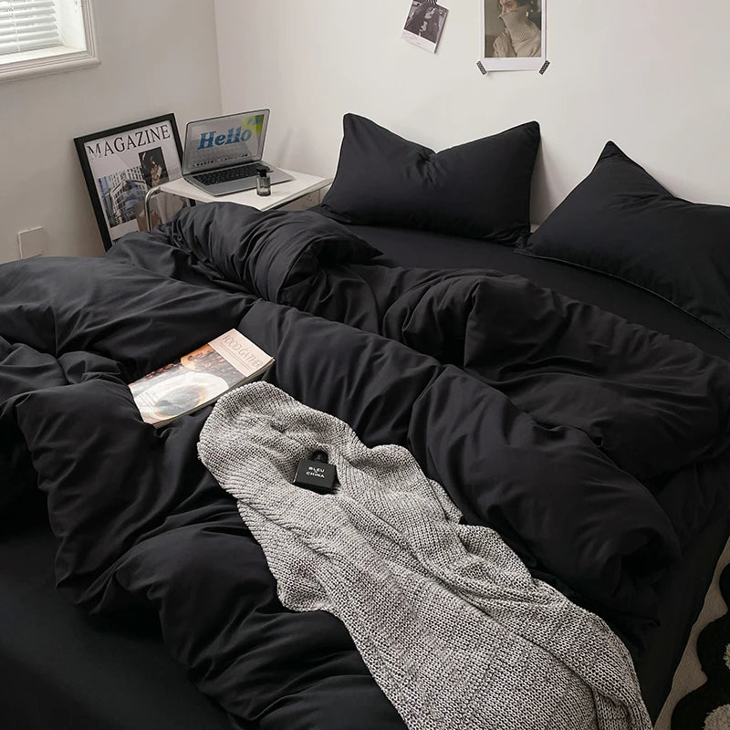 Bed with black bedding and a gray blanket, with a laptop and magazines on a nightstand.