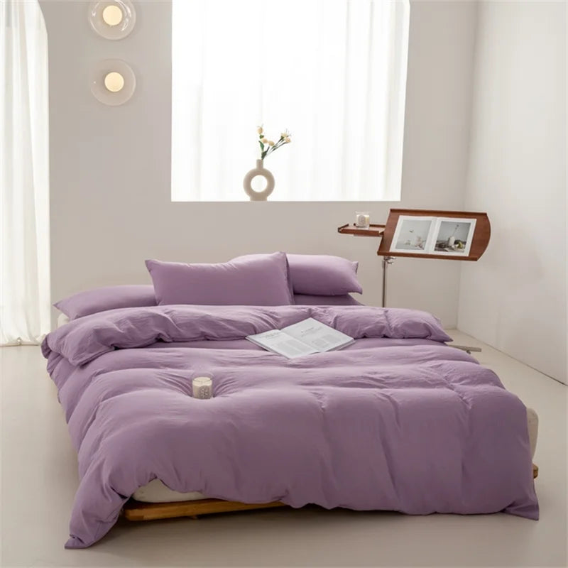 Purple bedding set on a bed in a minimalistic room with a window and side table.