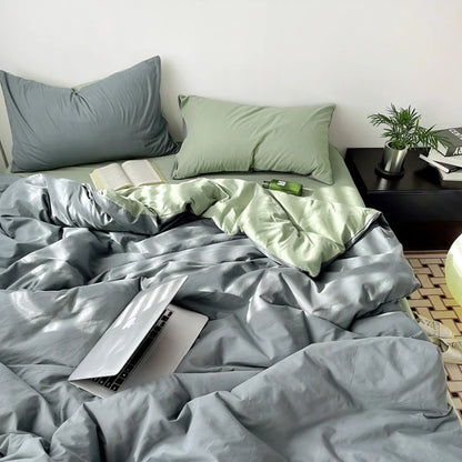 Bed with gray and green bedding, laptop on bed, nightstand with books and plant.