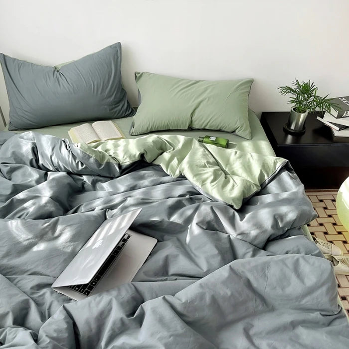 Bed with gray and green bedding, laptop on bed, nightstand with books and plant.