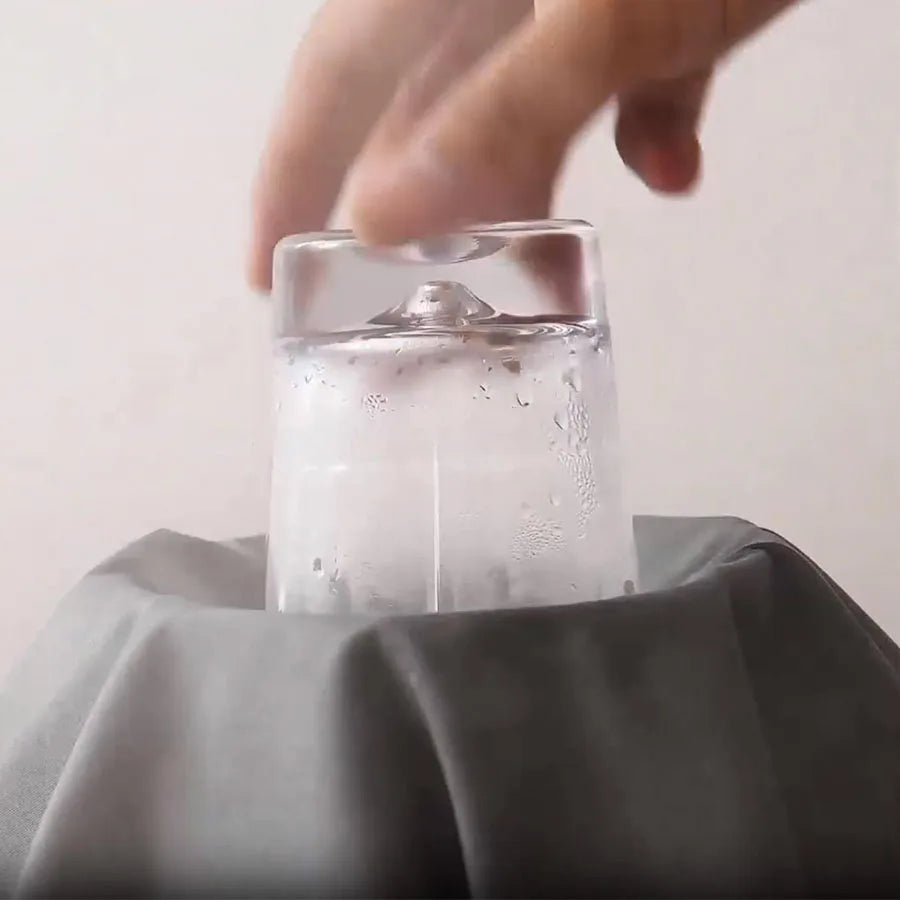 Clear glass of water with a hand reaching towards it on a gray surface.