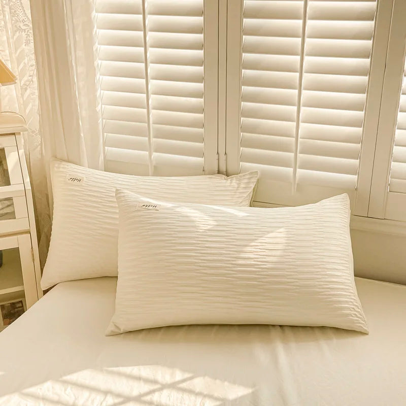 Two white pillows on a bed with light-colored walls and window blinds in the background.