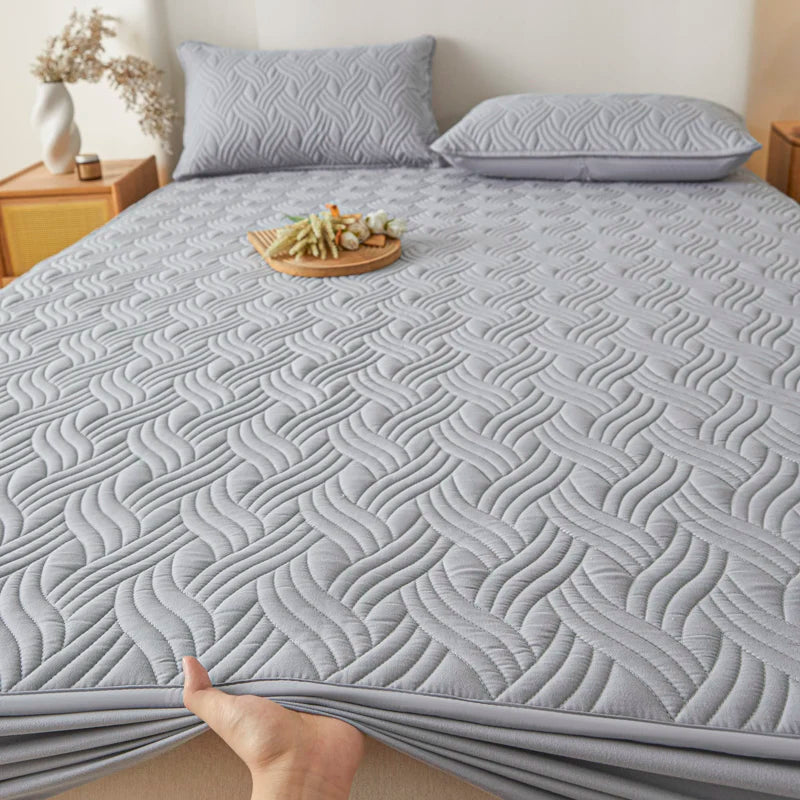 Gray quilted mattress cover on a bed with pillows and a small table in the background.