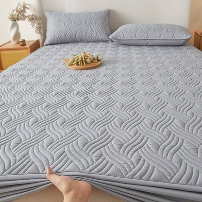 Gray quilted mattress cover on a bed with pillows and a small table in the background.