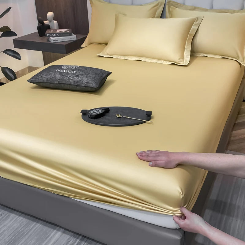 Person adjusting a gold satin sheet on a bed with a decorative pillow and small table in the background.